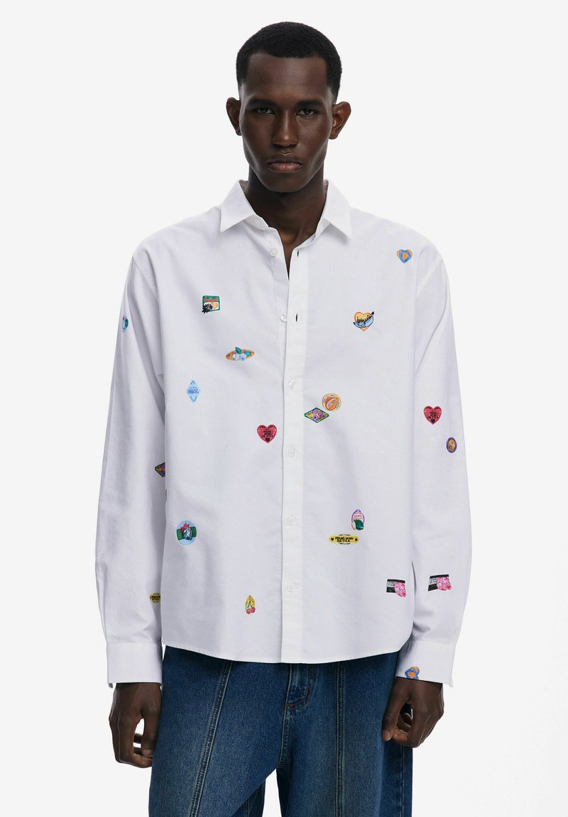 Camisa blanca de manga larga con cuello abotonado, que presenta coloridos parches bordados en varias formas y diseños distribuidos uniformemente por toda la tela.