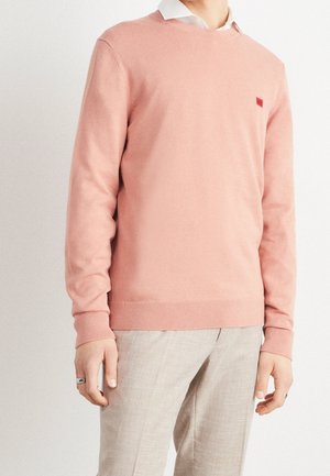 Person iført en lyserød crewneck-trøje over en hvid skjorte med krave og beige bukser, stående foran en ensfarvet baggrund.