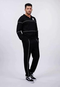 Ensemble de sport noir avec polo à manches longues orné de rayures blanches et de la lettre "B", associé à un pantalon assorti, complété par des baskets noires.