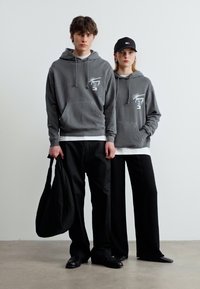 Δύο άτομα που φορούν ταιριαστά γκρι φούτερ με κουκούλα και μαύρο παντελόνι στέκονται μπροστά σε έναν λευκό τοίχο· ο ένας κρατά μια μεγάλη μαύρη τσάντα και ο άλλος φορά μαύρο καπέλο.