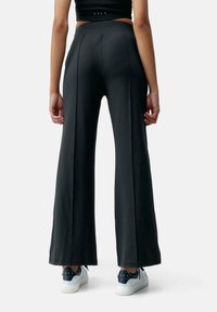 Pantaloni neri a gamba larga con una texture liscia, caratterizzati da una vita alta e dettagli plissettati. Abbinati a sneakers bianche, mostrano uno stile casual.