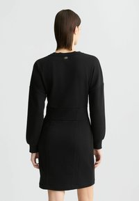 Čierna mikinová šaty s dlhými rukávmi, priliehavým pásom a rebrovanými manžetami. Na zadnom výstrihu sa nachádza dekoratívny logový detail.