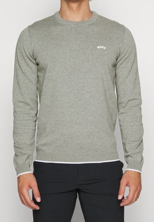 Mann trägt einen grauen Langarmpullover mit kleinem weißen "BOSS" Text auf der linken Brust und schwarzen Hosen vor einem einfarbigen Hintergrund.