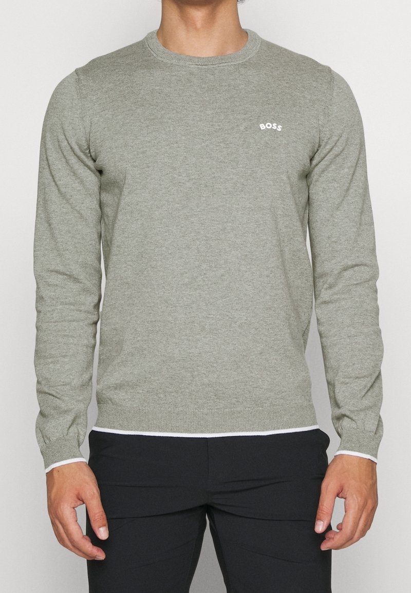 Homme portant un pull gris à manches longues avec un petit texte blanc "BOSS" sur la poitrine gauche et un pantalon noir sur un fond uni.