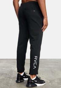 Zwarte sweatpants met een relaxte pasvorm, voorzien van een logo op het onderbeen en een klein achterzakje. Draagt met zwarte sportschoenen.