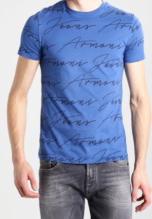 Homme portant un t-shirt bleu ajusté à manches courtes avec un texte noir en cursive "Armani Jeans" répété et un jean gris délavé.