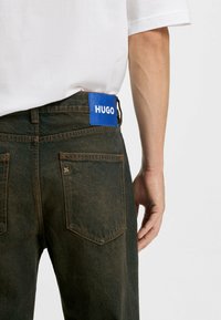 Vaqueros de denim de color verde oscuro con un acabado texturizado. Presentan una etiqueta azul con el logotipo blanco "HUGO" en la cintura. Dos bolsillos traseros.