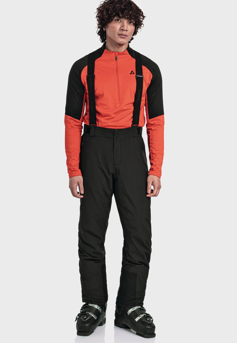 Mann in einem orange-schwarzen Langarmshirt, schwarzen Skihosen mit Hosenträgern und schwarzen Skischuhen, der vor einem schlichten Hintergrund steht.