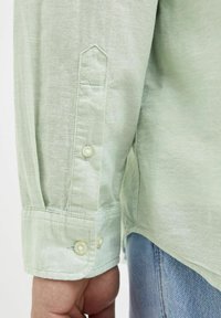 Camisa de manga larga verde claro hecha de una tela texturizada, que presenta un puño puntiagudo con dos botones y un dobladillo curvado.
