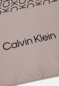 Sciarpa Calvin Klein in morbido tessuto beige chiaro, con un motivo ripetuto del logo "CK" e il nome del marchio in evidenza con caratteri neri e in grassetto.