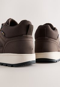 Paire de chaussures de sport marron avec semelles blanches, bande de roulement noire et languettes à l'arrière, vues d'un angle arrière bas.