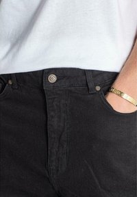 Pantalones de mezclilla negros con un corte clásico, que cuentan con detalles de botones de metal y bolsillos frontales, combinados con una camiseta blanca.