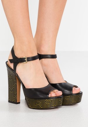 Czarne skórzane sandały na wysokim obcasie z odkrytymi palcami, regulowanym paskiem wokół kostki oraz złotym geometrycznym wzorem na masywnym obcasie.