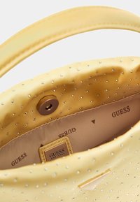 Gelbe Handtasche mit glatter Oberfläche, besetzt mit kleinen metallischen Akzenten. Innenraum mit beigem Futter, Markenzeichen und Magnetverschluss.