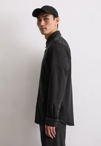 Man in een zwarte baseballcap, een zwart denim overhemd met knopen en zwarte jeans, staande in profiel tegen een effen lichte achtergrond.