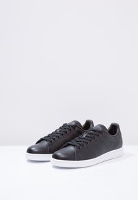 Pár černých kožených nízkých tenisek s bílými podrážkami a černými tkaničkami, umístěných vedle sebe na bílé ploše.