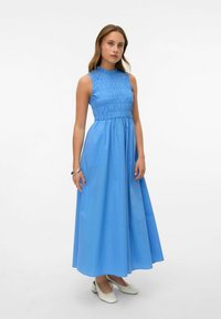 Modrá maxi šaty bez rukávů s nařasenou podprsenkou a vysokým límcem. Hladká tkanina, volná sukně, doplněná bílými botami.