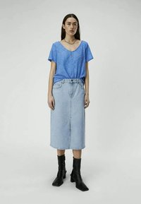 Woman standing wearing a blue short-sleeve blouse, light denim midi skirt, black ankle boots, and a gold chain necklace.