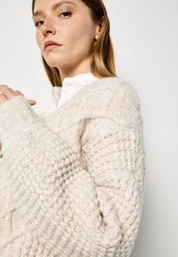 Grob gestrickter cremefarbener Pullover mit strukturiertem Muster und gerippten Bündchen. Das Model trägt ein weißes Hemd mit Kragen darunter.