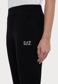 Svarta joggingsbyxor med en slät yta, med sidofickor och en vit EA7-logotyp på låret. Design med elastisk midja.