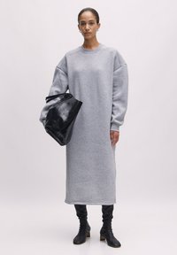 Graues, �übergroßes Sweatkleid mit langen Ärmeln, Rundhalsausschnitt und gerippten Bündchen. Kombiniert mit einer schwarzen, strukturierten Handtasche und kniehohen Stiefeln.