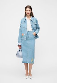 Kvinde står iført en lyseblå broderet denimjakke og nederdel, hvid bluse, hvide spidse sko og holder en sølvfarvet taske.