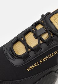Sneaker noire avec un dessus texturé noir, un panneau en maille dorée et des lacets contrastants. Marque "VERSACE JEANS COUTURE" en doré sur le côté.