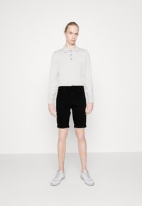Lys grå langermet polo med tre knapper, kombinert med svarte denimshorts. Modellen har på seg lys grå sneakers og står foran en hvit bakgrunn.
