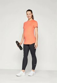 Mujer embarazada con camiseta naranja y leggings negros sosteniendo una botella de agua negra, de pie y sonriendo frente a un fondo liso.