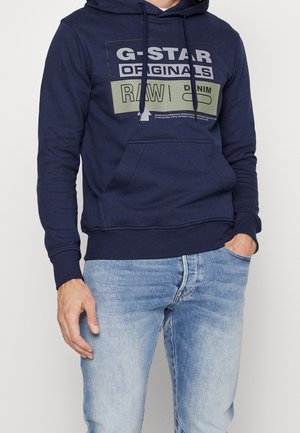 Mann trägt eine marineblaue G-Star Originals Kapuzenjacke mit Fronttasche und hellblaue Jeans, steht mit einer Hand in der Tasche.
