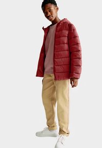 Chaqueta roja acolchada con costuras horizontales, sudadera gris debajo, pantalones beige con rayas laterales y zapatillas blancas.