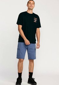 Black cotton t-shirt with front graphic, paired with blue cargo shorts featuring pockets and rolled cuffs. Worn with black sneakers and socks.