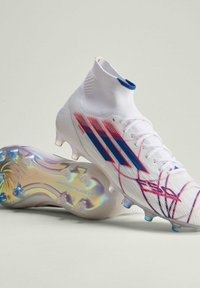 Λευκά ποδοσφαιρικά παπούτσια με μπλε και κόκκινες ρίγες, ψηλό περιλαίμιο αστραγάλου σαν κάλτσα, και ιριδίζουσα σόλα με καρφιά σχεδιασμένη για σκληρό έδαφος.