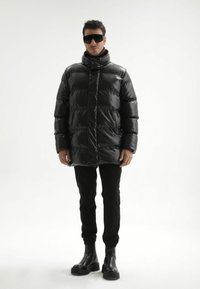 Zwarte donzen jas met een hoge kraag, voorzien van doorgestikte delen en zijzakken, gecombineerd met strakke zwarte broek en stevige zwarte laarzen.