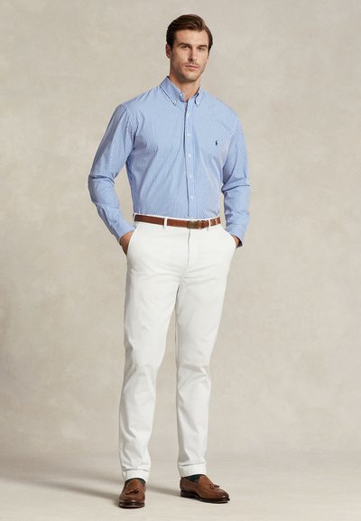 Mann iført blå og hvit stripet skjorte med knapper, hvite bukser, brunt belte og brune loafers med dusk, står med hendene i lomma.