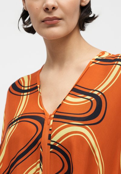 Γυναίκα με πορτοκαλί τοπ με κίτρινες και μαύρες καμπυλωτές γραμμές, με φερμουάρ μπροστά και γιακά σε σχήμα V.