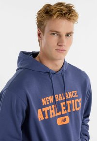 Felpa con cappuccio blu navy decorata con la scritta "NEW BALANCE ATHLETICS" in arancione. Tessuto morbido, con cordino regolabile e tasca anteriore a marsupio.