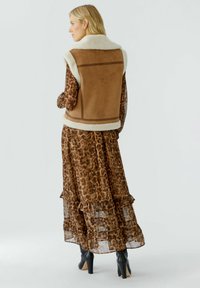 Frau in einem durchsichtigen Kleid mit Leopardenmuster und einer beigefarbenen ärmellosen Schafwollweste, trägt schwarze Absatz-Stiefeletten und steht seitlich vor einem weißen Hintergrund.