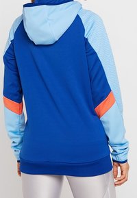 Blå sportig hoodie med ljusblå huva och ärmdekorationer, har orange och ljusblå färgblockering, med en texturerad design på ärmarna.