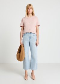 Hellrosa kurzärmeliges T-Shirt mit kleinen Verzierungen, kombiniert mit hellblauen, weit geschnittenen Jeans und einer braunen Wildlederhandtasche. Beige spitz zulaufende High Heels.