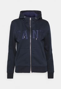 Sudadera con cremallera de color azul marino hecha de tela suave. Presenta una capucha con cordón, bolsillos frontales y un logo bordado de "GANT" en un color a juego.