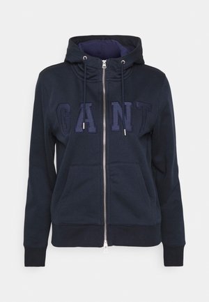 Sudadera con cremallera de color azul marino hecha de tela suave. Presenta una capucha con cordón, bolsillos frontales y un logo bordado de "GANT" en un color a juego.