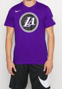 T-shirt Nike violet à manches courtes avec un logo noir circulaire portant "LA" en blanc et violet, associé à un short de sport noir avec des accents blancs.