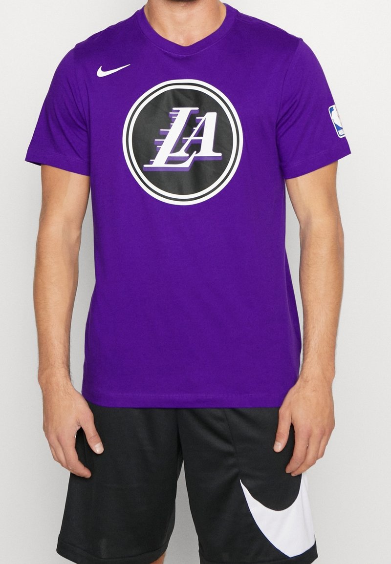 T-shirt Nike violet à manches courtes avec un logo noir circulaire portant "LA" en blanc et violet, associé à un short de sport noir avec des accents blancs.