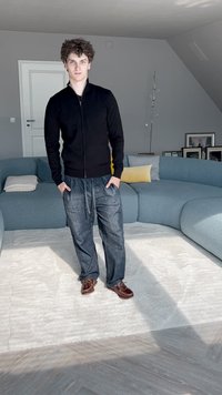 Svart zip-up jacka, lösa blå jeans och bruna skor. Står på en ljus matta i ett rum med en böjd blå soffa och dämpad belysning.