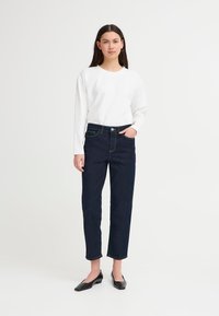 Mujer con cabello largo y oscuro, vistiendo una camisa blanca de manga larga, jeans oscuros y zapatos planos negros de punta, de pie frente a un fondo blanco liso.