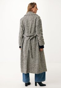 Cappotto lungo in motivo houndstooth grigio e nero, con vita cinturata, ampio colletto e tessuto testurizzato. Abbinato a jeans a gamba larga e stivali.