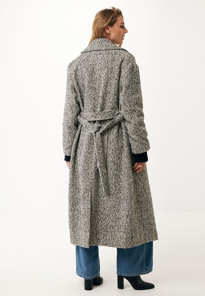 Lange, grijs-zwart houndstooth-gepatroneerde jas met een riem in de taille, grote kraag en textuurstof. Gestyled met wijde jeans en laarzen.