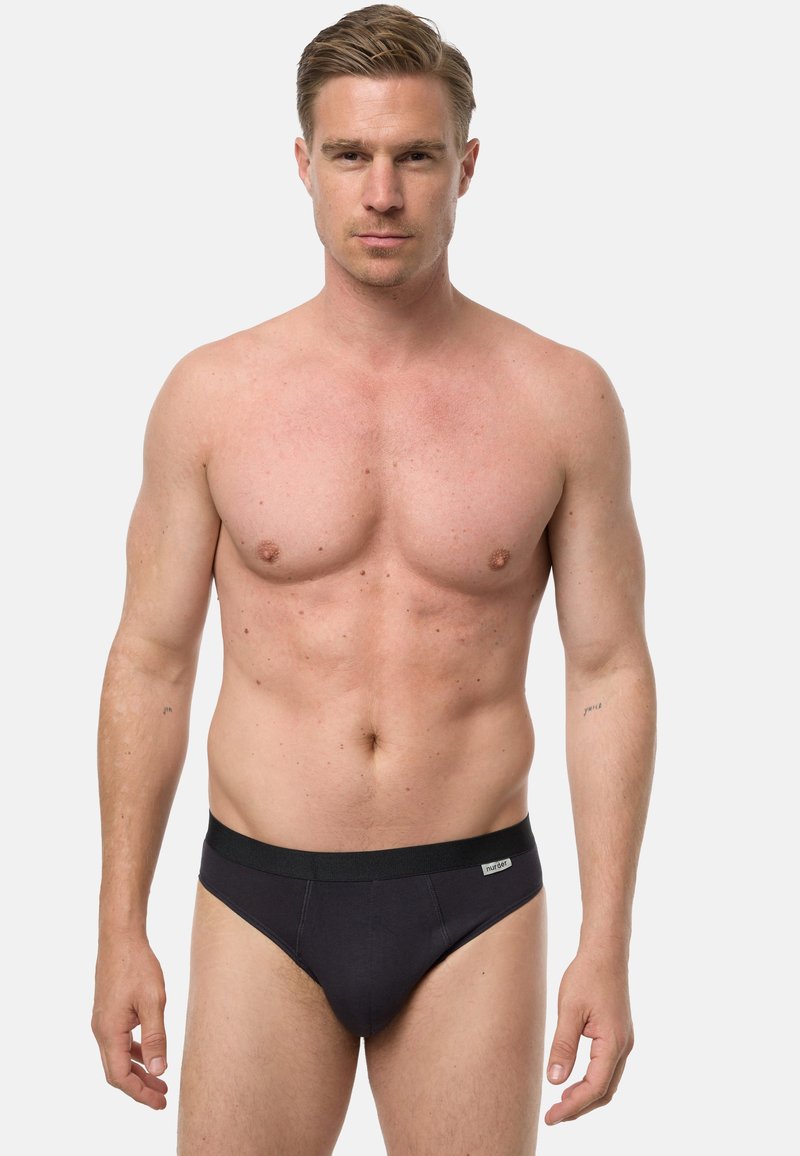 Homme torse nu aux cheveux courts et clairs portant un slip noir, debout face à l'avant sur un fond clair uni.