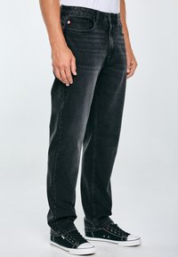 Jeans in denim nero con taglio dritto, caratterizzati da un design classico a cinque tasche e da un piccolo etichetta con logo sulla tasca anteriore. Indossati con sneakers nere.